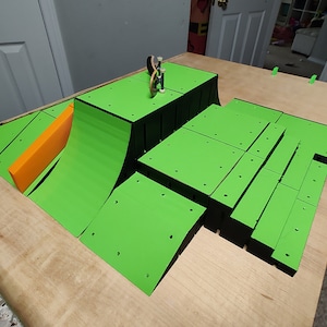 May include: A green and black fingerboard ramp set on a wooden surface. The ramp features a curved ramp, a flat platform, and a variety of other obstacles. A small fingerboard is on the ramp.