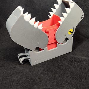 May include: A grey and red 3D printed dinosaur-shaped dice tower with a yellow eye. The tower has a hinged jaw that opens to reveal a red dice tray.
