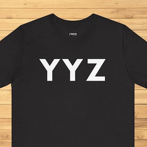 May include: Black t-shirt with the white letters "YYZ" across the chest. The bottom of the image shows the text "6 Color Variations". The shirt is displayed on a wooden surface.