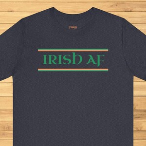 Könnte beinhalten: Marineblaues T-Shirt mit dem Text "IRISH AF" in Grün, mit grünen, weißen und orangefarbenen Streifen. Das T-Shirt wird auf einer Holzoberfläche präsentiert.
