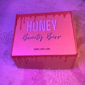 May include: A pink box with dripping pink paint on the sides. The box has the words "HONEY Beauty Barr" in white text and the social media handle "@HONEY_BEAUTY_BARR" in smaller white text.
