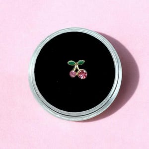 May include: A small, cherry-shaped stud earring with pink gemstones and green leaves, set in a gold-colored metal. The earring is displayed in a clear, round container with a black interior, set against a pink background.