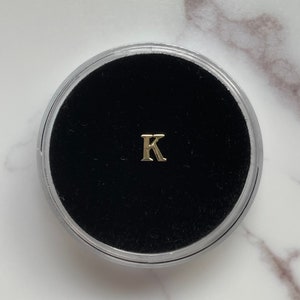 May include: A small, gold-colored letter "K" charm displayed in a clear, round container with a black velvet interior. The charm is centered, and the container is set against a white and gray marble surface.