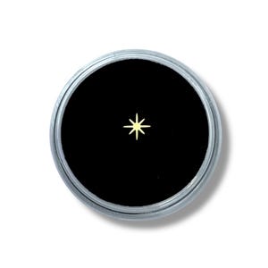 Puede incluir: Un pequeño objeto en forma de estrella de color dorado está centrado sobre un fondo negro dentro de un recipiente circular plateado. La estrella tiene ocho puntas y es el punto focal de la imagen.