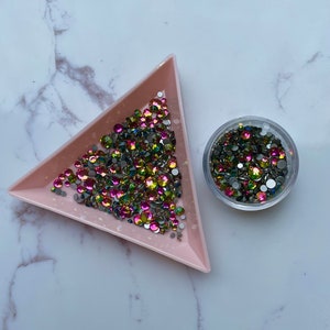May include: A pink triangular tray filled with iridescent green, pink, and silver rhinestones. A clear plastic container filled with the same rhinestones is next to the tray.