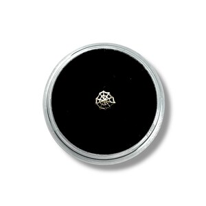 Puede incluir: Un pequeño piercing de nariz dorado en forma de telaraña. El piercing está centrado en un fondo de terciopelo negro, encerrado en un recipiente redondo de color plateado. El fondo es blanco.