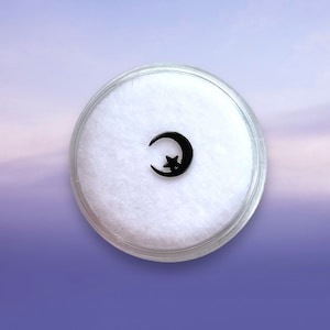 May include: A small black crescent moon charm with a star inside, displayed on a white background in a clear plastic container.