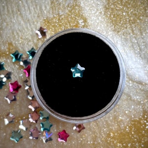 May include: A small, iridescent star-shaped gem sits in a black velvet-lined container. Scattered around the container are more star-shaped gems in various colors, including pink, blue, and silver. The background is a textured, gold-colored surface.