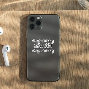 May include: A black iPhone with a white sticker that says "Maybe Today SATAN Maybe Today" on a wooden surface with two white wireless earbuds.