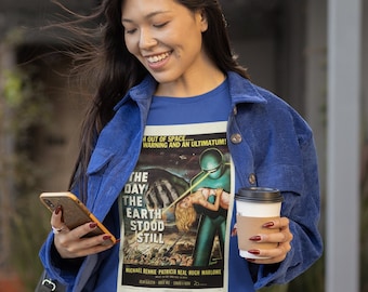 Könnte beinhalten: Eine Frau trägt ein blaues Shirt mit einem Vintage-Filmplakat-Design, das einen grünen Außerirdischen und den Text "Der Tag, an dem die Erde stillstand" zeigt. Sie hält ein Smartphone in der einen Hand und eine Tasse Kaffee in der anderen.