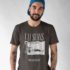 Könnte beinhalten: Ein dunkelgraues T-Shirt mit einer Grafik im Vintage-Stil, die eine Frau zeigt, die einen CT-Scan erhält. Die Grafik lautet "Cat Scans of the Future" und "Hold Your Breath".