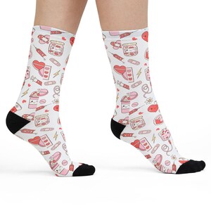 May include: White socks with a repeating pattern of pink and red hearts, jars, bandages, and other cute illustrations. The socks have black heels and toes. The socks are knee-high.