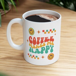 May include: White ceramic coffee mug with a colorful retro design. The mug has the text "Coffee Makes Me Happy" with smiley faces and geometric shapes.