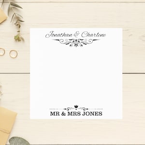May include: A white card with black text that reads "Jonathan & Charlene" with a decorative flourish above the text and "MR & MRS JONES" with a decorative flourish below the text. The card is on a white wooden surface.