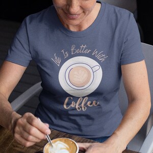 Może przedstawiać: Kobieta w granatowym t-shircie z napisem "Life is better with coffee" i grafiką przedstawiającą filiżankę kawy. Siedzi przy stole z filiżanką kawy przed sobą.
