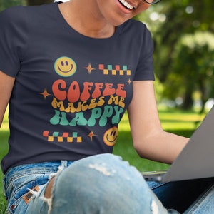Puede incluir: Una mujer con una camiseta azul oscuro con el texto "Coffee Makes Me Happy" en una fuente retro y caritas sonrientes de colores. Está sentada en un muro de piedra con un portátil y una taza de café para llevar.