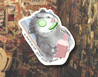 Op de afbeelding: Een sticker met een cartoon opossum met komkommerschijfjes op zijn ogen, die een roze fles vasthoudt. De tekst "moisturize your flesh prison" staat op de sticker. De opossum is grijs en wit, met roze accenten.
