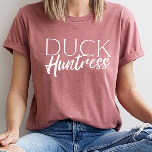 May include: A woman wearing a dusty rose t-shirt with the text "DUCK Huntress" in white lettering. She is wearing blue jeans with a distressed look.