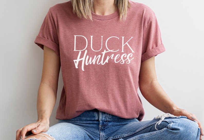 May include: A woman wearing a dusty rose t-shirt with the text "DUCK Huntress" in white lettering. She is wearing blue jeans with a distressed look.