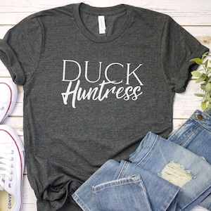 May include: A dark gray t-shirt with the text "DUCK Huntress" in white lettering. The shirt is laid flat on a white wooden surface with a pair of white sneakers and a pair of blue jeans.