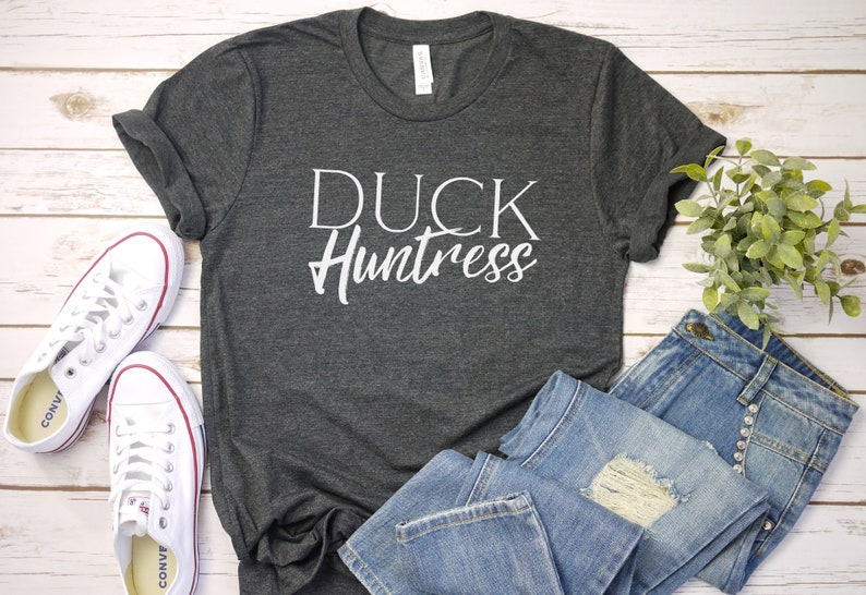 May include: A dark gray t-shirt with the text "DUCK Huntress" in white lettering. The shirt is laid flat on a white wooden surface with a pair of white sneakers and a pair of blue jeans.