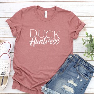 May include: A dusty rose t-shirt with the text "DUCK Huntress" in white lettering. The t-shirt is laid out on a white wooden surface with a pair of white sneakers and denim shorts.