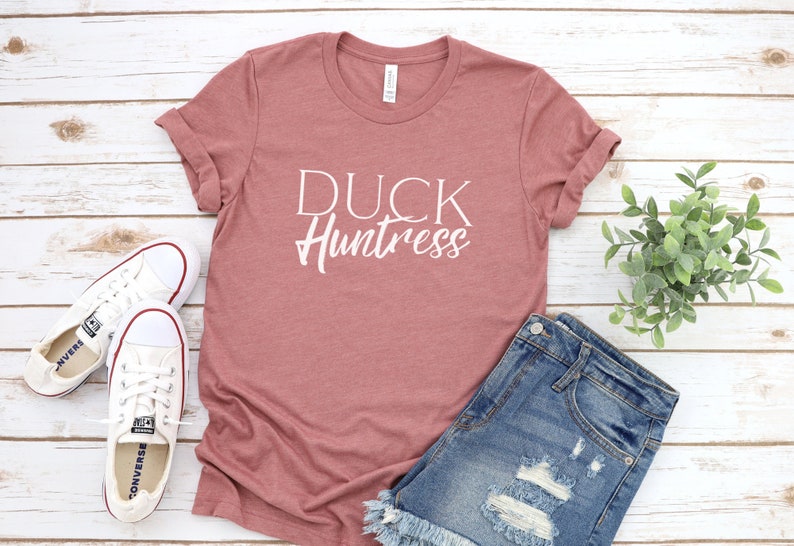 May include: A dusty rose t-shirt with the text "DUCK Huntress" in white lettering. The t-shirt is laid out on a white wooden surface with a pair of white sneakers and denim shorts.