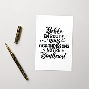 May include: A white card with black calligraphy that reads "B&eacute;b&eacute; en route. nous agrandissons notre bonheur!"  The card is on a white surface with two pens.