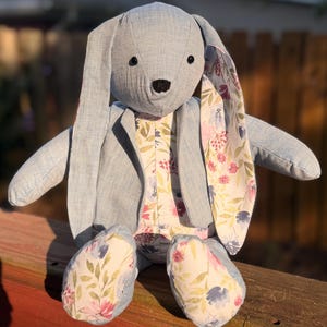 Pode incluir: Um coelho de pelúcia feito de tecido azul claro com orelhas longas. O interior das orelhas, o peito e os pés apresentam um padrão floral com folhas e flores rosas, azuis e verdes. O coelho tem dois pequenos olhos de contas pretas.