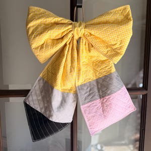 Op de afbeelding: Een handgemaakte, gewatteerde strik in de vorm van een potlood, hangend aan een deur. De strik heeft gele gingham stof voor de strik, met grijze, roze en zwarte secties die het lichaam en de punt van het potlood voorstellen. De strik wordt opgehangen door een natuurlijk touw.