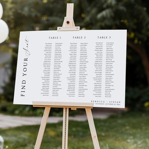 May include: A white rectangular wedding seating chart sign on a light wood easel. The sign features the text "FIND YOUR SEAT" vertically on the left, three columns labelled "TABLE 1", "TABLE 2", and "TABLE 3" with guest names, and "REBECCA + STEFAN" at the bottom.
