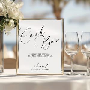 May include: A rectangular sign with a gold frame featuring the black text Cash Bar, the phrase DRINKS ARE ON YOU, THE MEMORIES ARE ON US, the word cheers, and the names REBECCA + STEFAN printed on a white background in a mix of script and serif fonts.