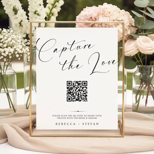 May include: A rectangular sign in a thin gold frame featuring the black text Capture the Love, a central QR code, and the names Rebecca + Stefan. The sign includes the instruction Please scan the QR code to share your photos with the bride & groom.