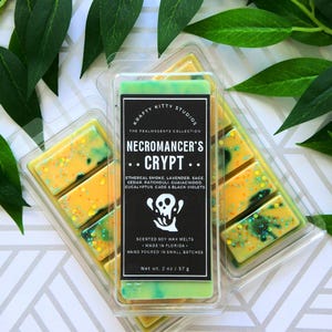 May include: A green, yellow, and black wax melt with a skull graphic and the text "Necromancer's Crypt". The wax melt is in a clear plastic container and is labeled "Krafty Kitty Studios" and "The Realmscents Collection". The wax melt is scented with ethereal smoke, lavender, sage, cedar, patchouli, guaiacwood, eucalyptus, cade, and black violets. The wax melt is 2 ounces.