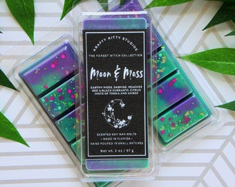 Könnte beinhalten: Zwei durchsichtige Kunststoffbehälter gefüllt mit violetten und grünen Duftwachs-Melts aus Sojawachs. Das Etikett trägt die Aufschrift Krafty Kitty Studios, The Forest Witch Collection, Moon & Moss, Earthy Moss, Jasmine, Peaches, Red & Black Currants, Citrus, Hints of Tonka and Amber, Net wt. 57 g.