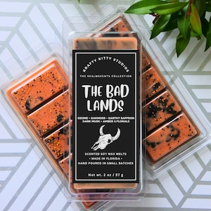 May include: Soy wax melts in a clear plastic container. The melts are orange with black speckles. The label reads "The Bad Lands" with scent notes and a skull graphic. Net wt. 2 oz / 57 g.