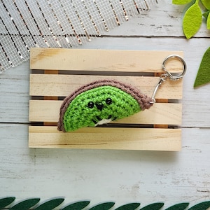 Puede incluir: Un llavero de kiwi de punto de crochet verde y marrón con una cara sonriente. El llavero está unido a un llavero plateado.