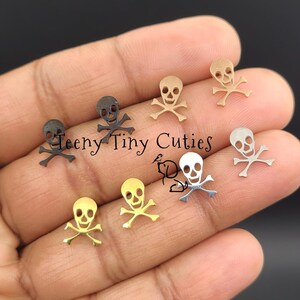 May include: A collection of seven small skull and crossbones charms in various metallic finishes including gold, silver, rose gold, and black. The charms are arranged in a cluster and feature the text Teeny Tiny Cuties overlaid in the centre.
