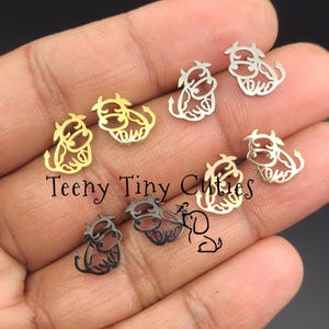 May include: A collection of small, flat, metal cow-shaped stud earrings in gold, silver, and black finishes. Each earring features a cutout design of a cow with horns and a tail. The text Teeny Tiny Cuties is printed in black across the centre.