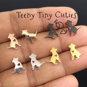 May include: A collection of small dog-shaped stud earrings in silver, gold, and black metal finishes. Each earring features a cutout heart shape on the dog's body. The text Teeny Tiny Cuties is printed at the top of the image above the earrings.