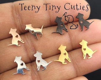 Pode incluir: Uma coleção de pequenos brincos em forma de cão com acabamentos em metal prateado, dourado e preto. Cada brinco apresenta um recorte em forma de coração no corpo do cão. O texto Teeny Tiny Cuties está impresso no topo da imagem.