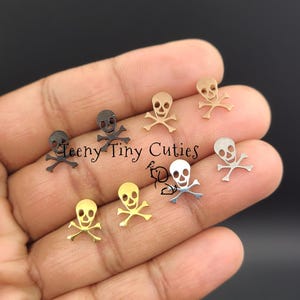 May include: A collection of small skull and crossbones charms in various metallic finishes, including gold, silver, and black. The charms are arranged on a hand, with the text "Teeny Tiny Cuties" visible.