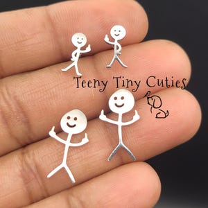 Peut inclure: Quatre boucles d'oreilles en forme de bonhomme allumette argenté avec des visages souriants et des doigts d'honneur levés. Le texte Teeny Tiny Cuties et une petite icône de chien au trait sont visibles au centre de ces bijoux métalliques.
