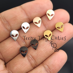 May include: A collection of small alien-shaped charms in various metallic finishes, including silver, gold, and black. The charms feature a minimalist design with large, oval eyes and a small chin. The text "Teeny Tiny Cuties" is visible.