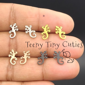May include: Four pairs of small lizard-shaped stud earrings in silver, gold, rose gold, and black metal finishes. The text Teeny Tiny Cuties is printed in black script in the center, accompanied by a small line drawing of a dog and a bird.