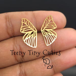 May include: A pair of gold-colored stud earrings shaped like butterfly wings with an intricate openwork vein pattern. The text Teeny Tiny Cuties is printed below the earrings, accompanied by a small line drawing of a dog with a bird.