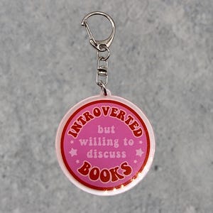 May include: A pink circular keyring with the text "INTROVERTED but willing to discuss BOOKS" in red and white. It has a silver clasp and chain. The background is a light grey.