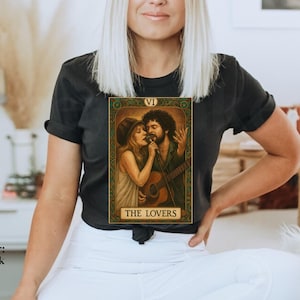 May include: Black t-shirt featuring a tarot card design with the text "THE LOVERS." The card depicts a couple with a guitar. The shirt is worn by a person. The color of the shirt is black.