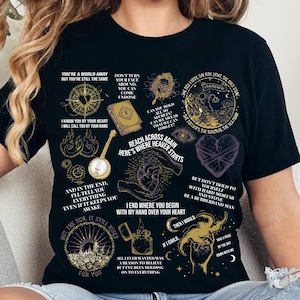 May include: A black short-sleeve t-shirt featuring a collage of gold and purple graphic illustrations including a compass, a book, a banjo, a heart, and a lighter, accompanied by various lines of text such as "Reach across again here's where heaven starts."