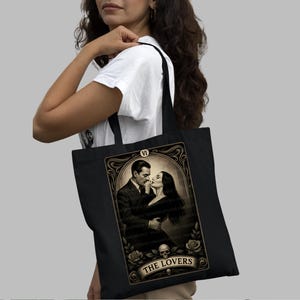 Black canvas tote bag featuring “The Lovers” tarot-inspired design inspired by Morticia and Gomez Addams. Perfect gothic romance gift for Addams Family fans, Halloween lovers, and dark aesthetic admirers. Handmade by House of Lotus Co, this high-quality tote celebrates eternal love, passion, and mystery with vintage-style tarot artwork in sepia tones.